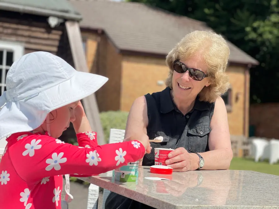 A grandmother and a young child in a sun hat sharing tubs of ice cream at an outdoor table in the café garden