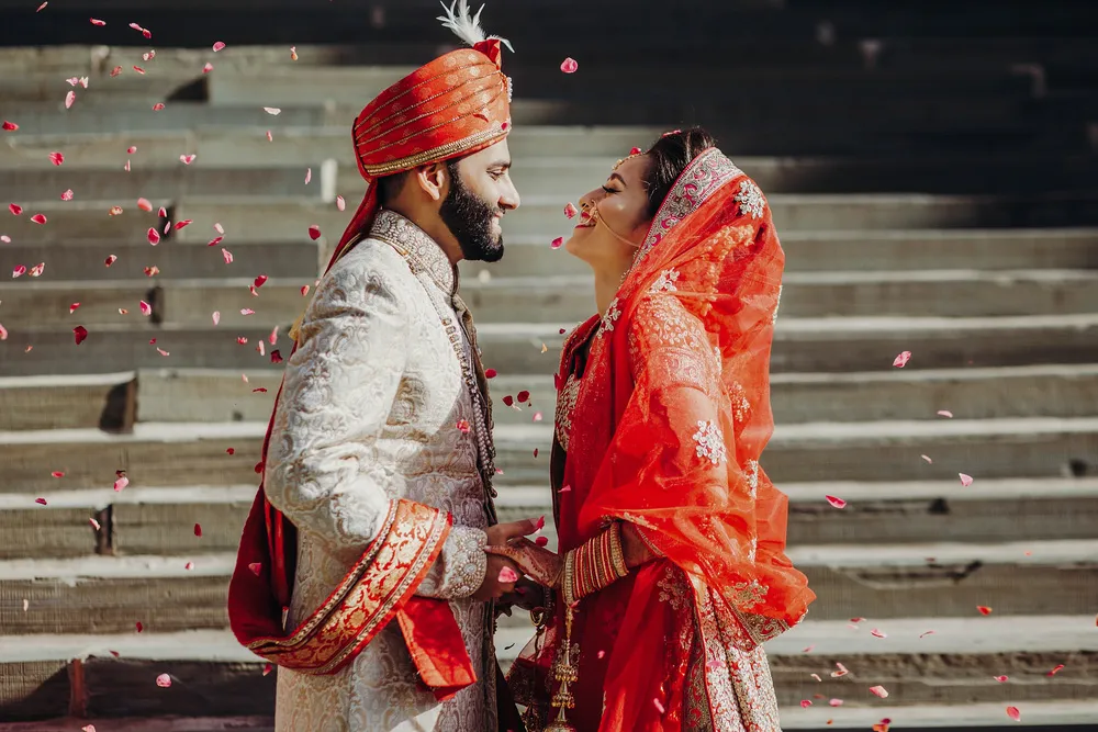 An Indian bride and groom in traditional dress facing one another as rose petals fall