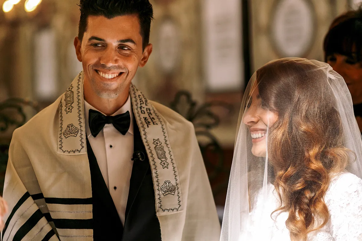 A smiling Jewish groom in a tallit beside his veiled bride during the ceremony