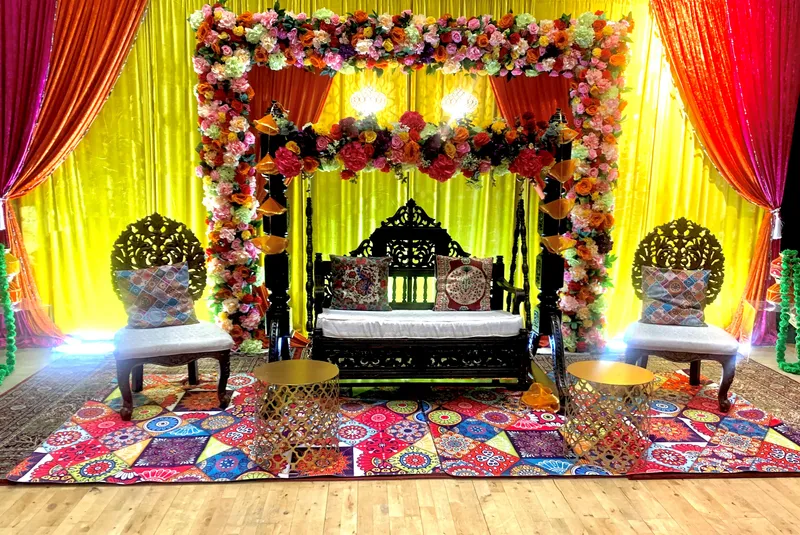 A colourful mehndi stage at GXCA with a carved wooden swing, floral garlands and bright drapes