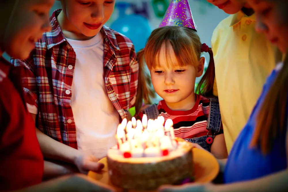 Children in party hats gathered around a birthday cake lit with candles
