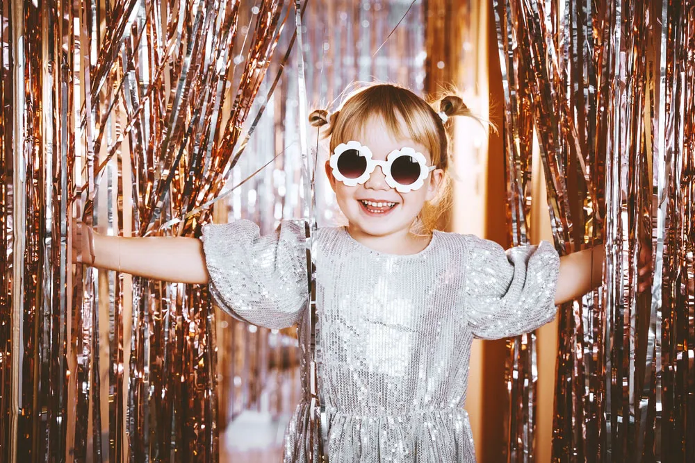 A young girl in a sequin dress and flower sunglasses posing at a party backdrop